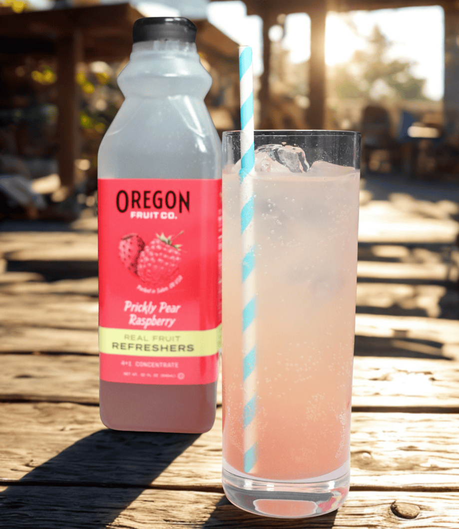Clear glass of pink beverage in front of a bottle of Prickly Pear Raspberry Refresher on a picnic table