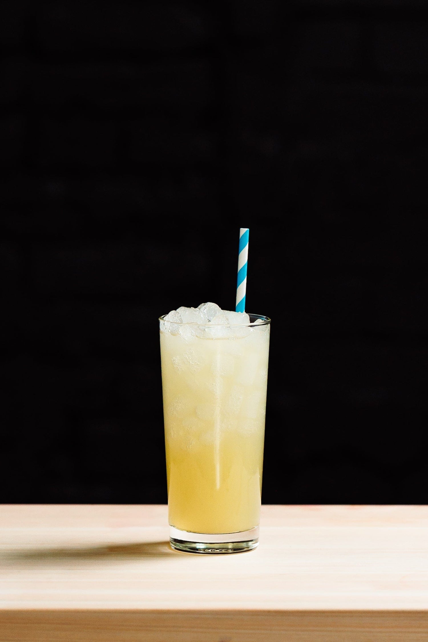 glass of ice and yellow refresher with a striped straw in front of black background