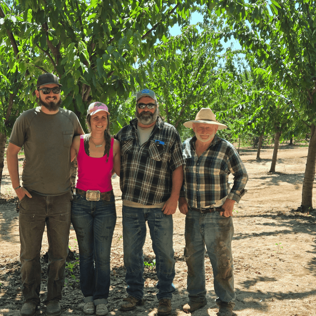 Four members of a farming family stands in their cherry orchard. A tall young man in a green shirt stands next to a young woman in a bright red shirt, who is next to an older man in a plaid button down, who is next to a senior man in a matching plaid
