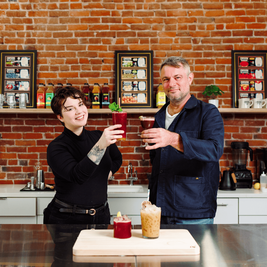A man and woman stand behind a stainless steel bar showing off their drinks