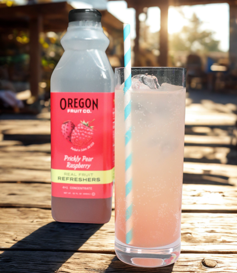 Clear glass of pink beverage in front of a bottle of Prickly Pear Raspberry Refresher on a picnic table