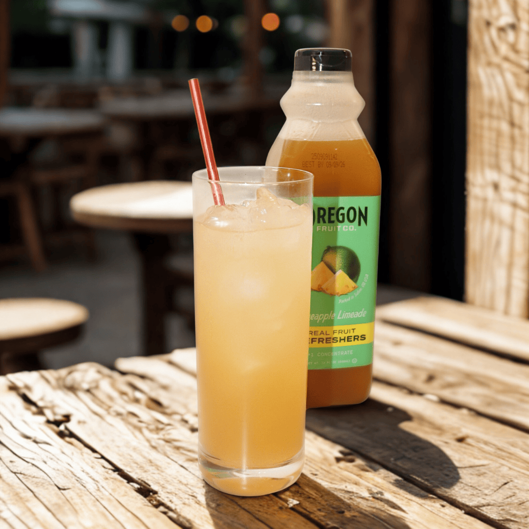 Clear glass of yellow beverage in front of a bottle of Pineapple Lemonade Refresher on a picnic table