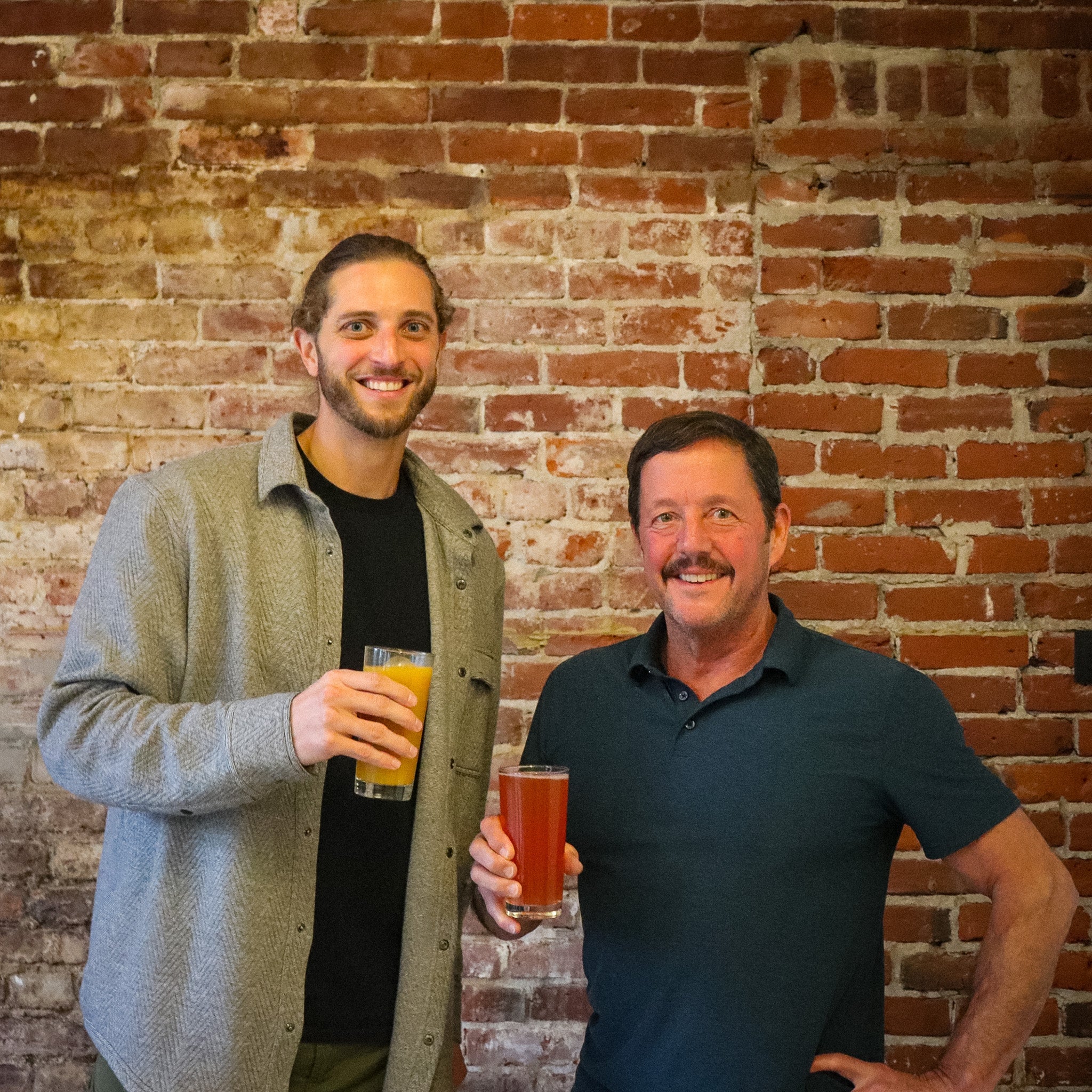 Two men stand in front of a brick wall holding glasses of fruited beer