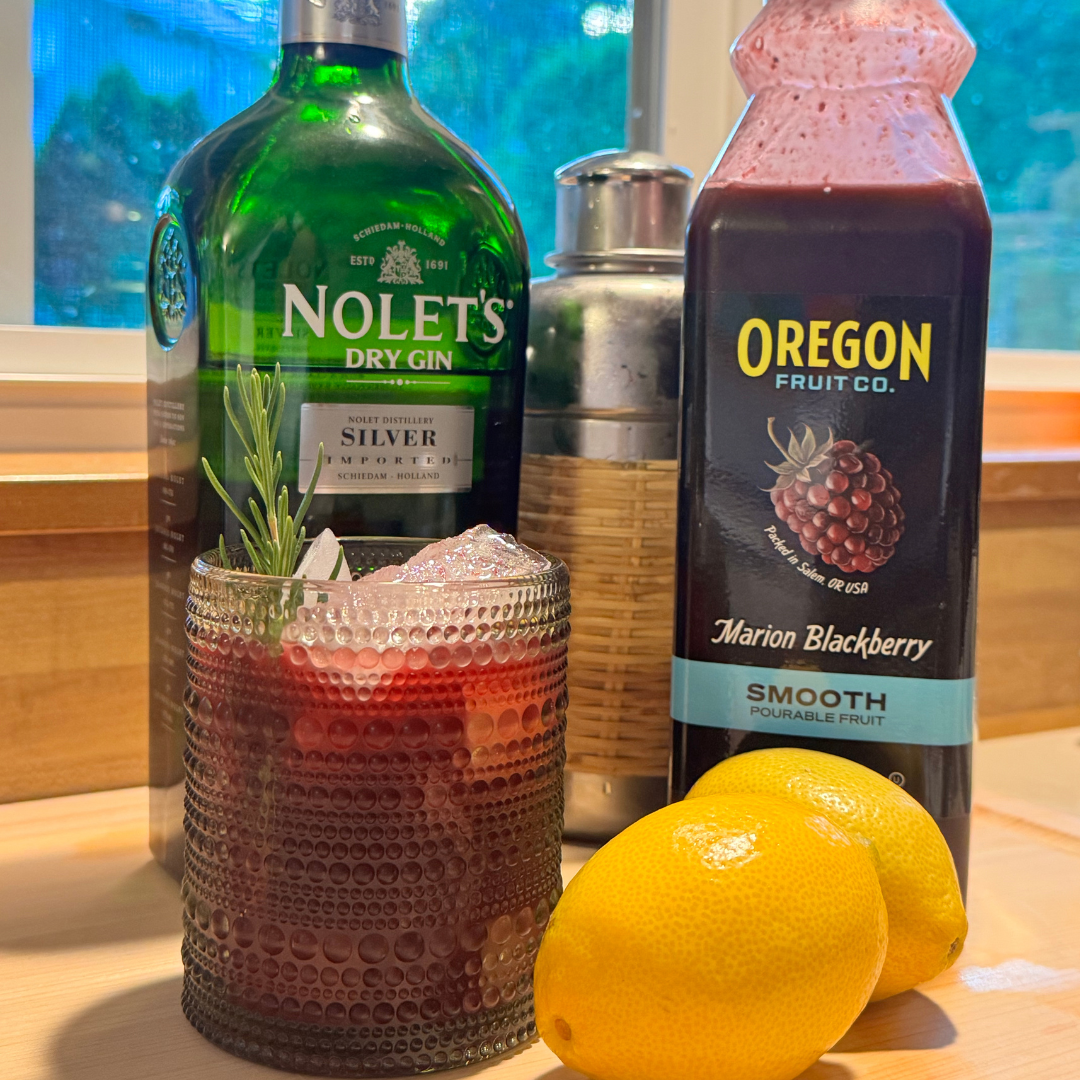 Textured glass with ice and red liquid next to lemons, shaker, bottle of gin and bottle of marionberry puree