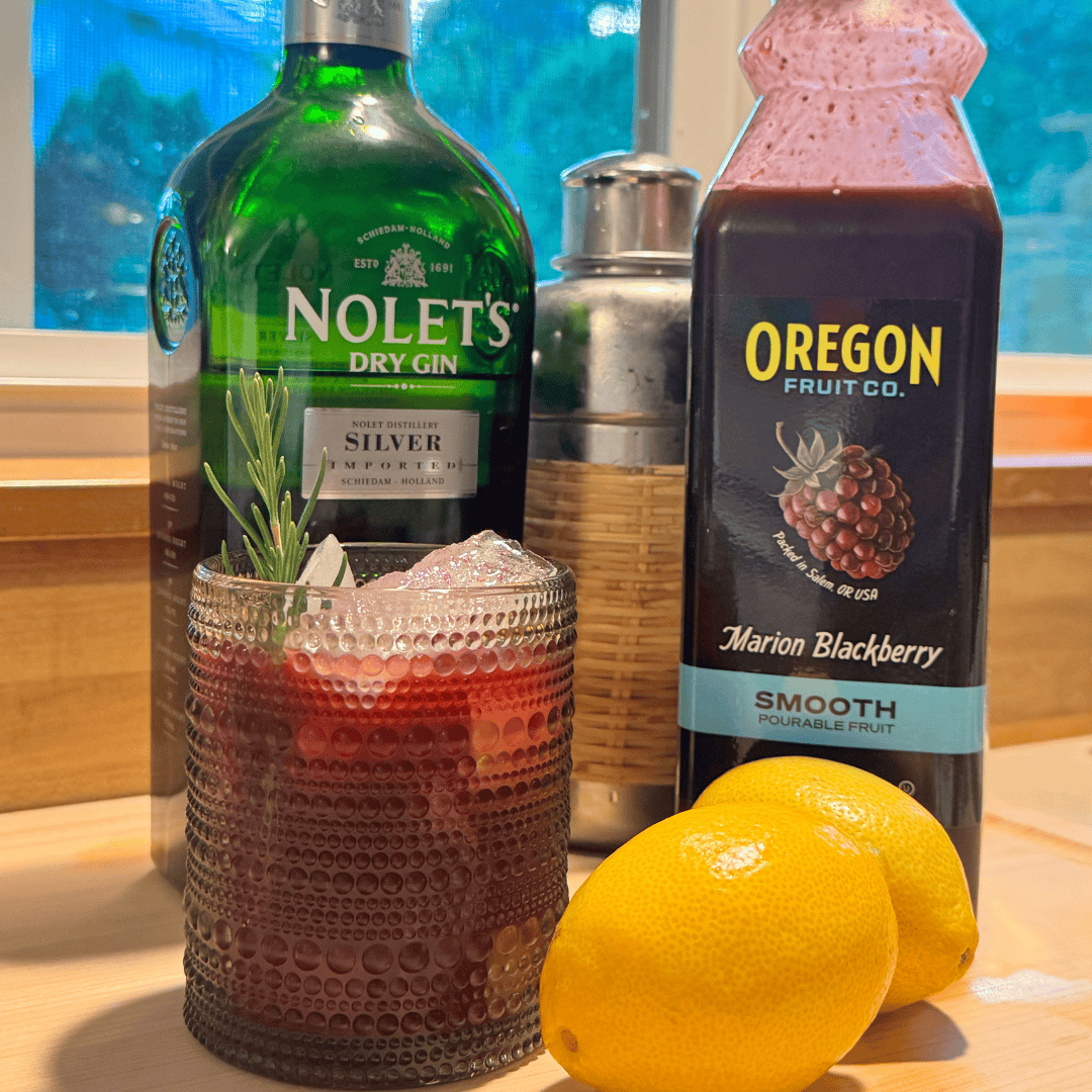 Textured glass with ice and red liquid next to lemons, shaker, bottle of gin and bottle of marionberry puree
