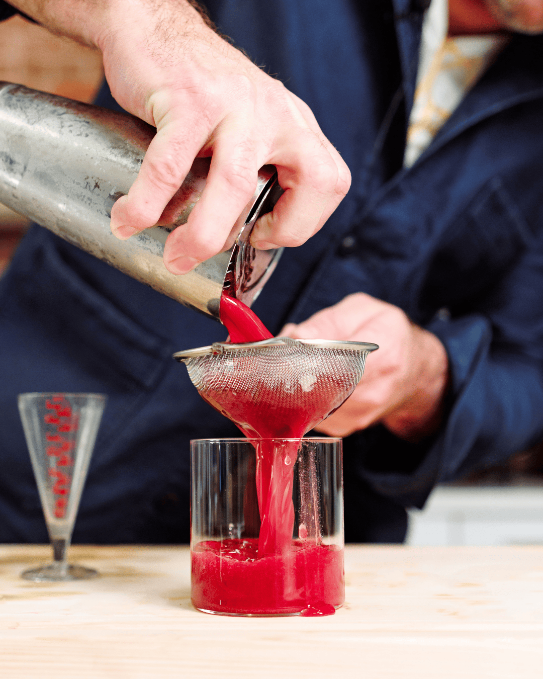 a bartender strains a bright red drink from a stainless steel shaker into a cocktail glass