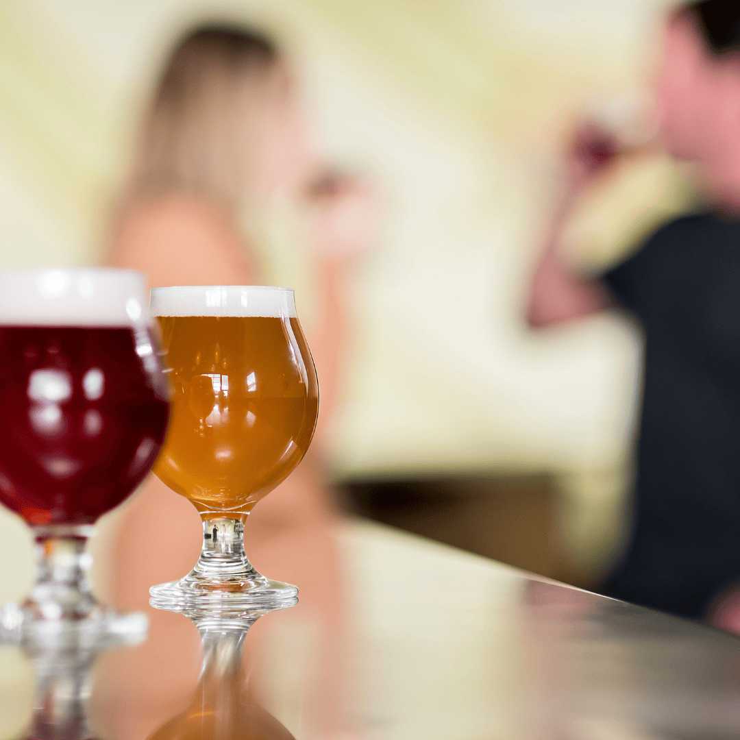 two glasses of beer sit on a bar while people drink in the background
